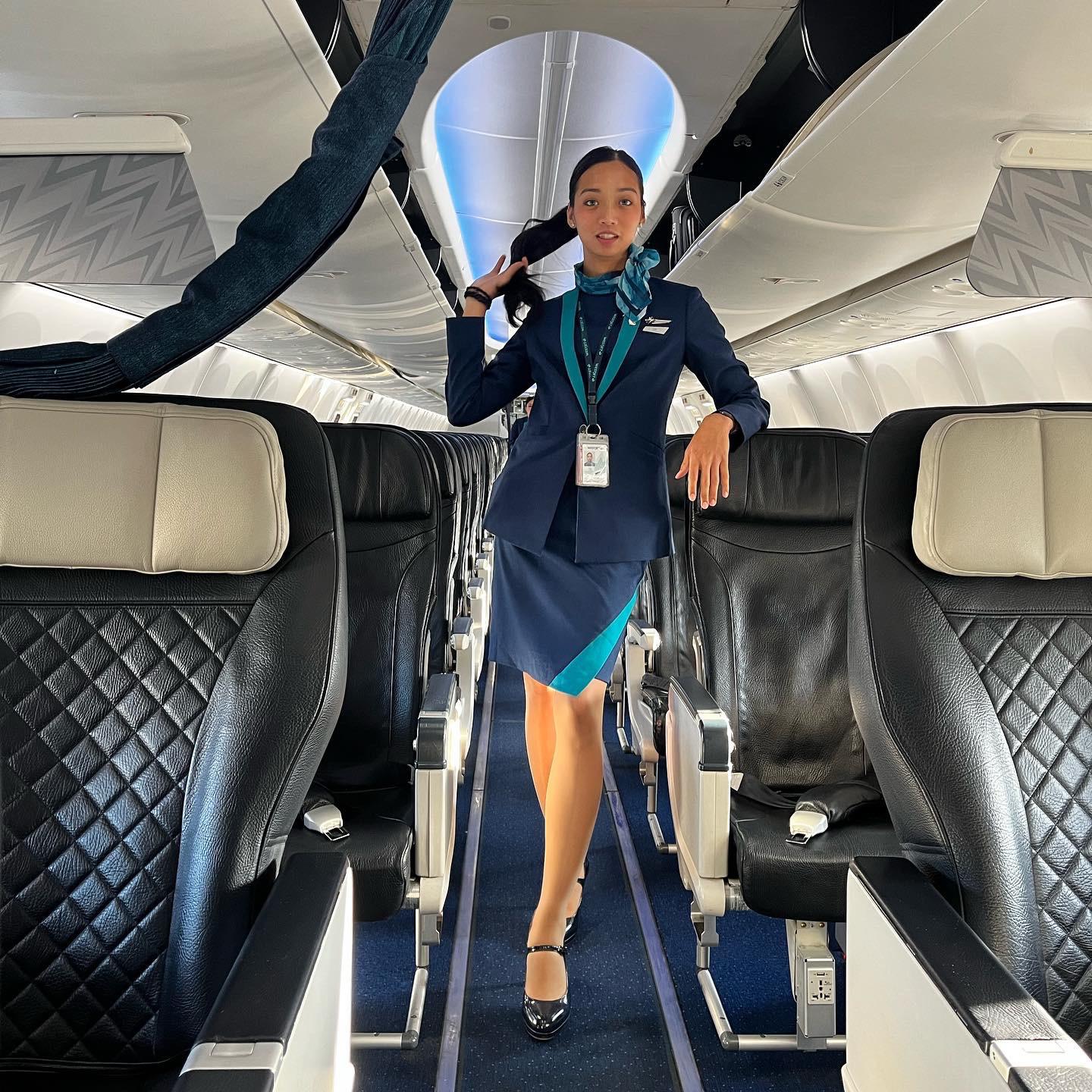 A flight attendant in a navy uniform stands confidently in the aisle of an airplane cabin, holding her hair. The aircraft seats are empty and the cabin is well-lit.