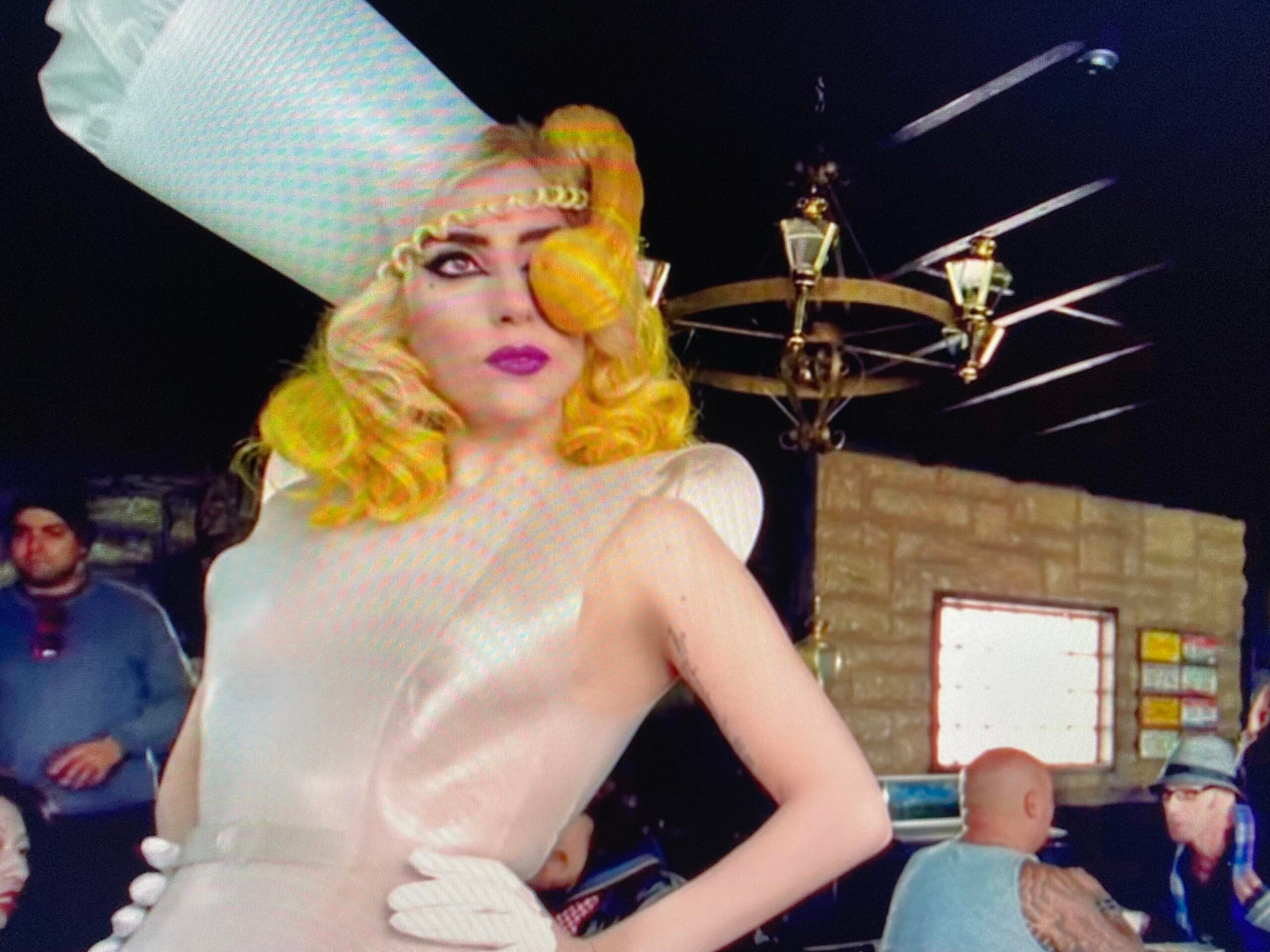 A person in a dramatic cream-colored dress, tall sculptural hat, and yellow hair poses confidently indoors. Several people are seated in the background under ornate lights in a casual setting.