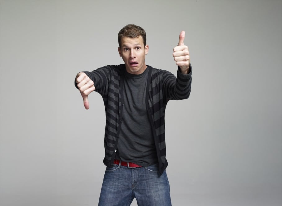 A man wearing a striped sweater and jeans stands against a plain background, giving a thumbs-up with one hand and a thumbs-down with the other, with a confused or uncertain expression on his face.