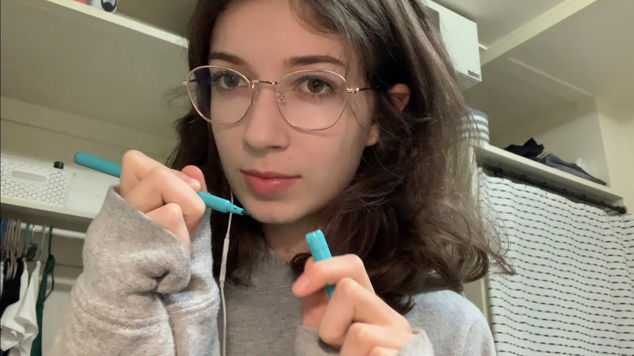 A young woman with shoulder-length brown hair and glasses holds a blue pen near her face. She wears a gray sweater with sleeves covering her hands and stands in a room with shelves and hanging clothes in the background.