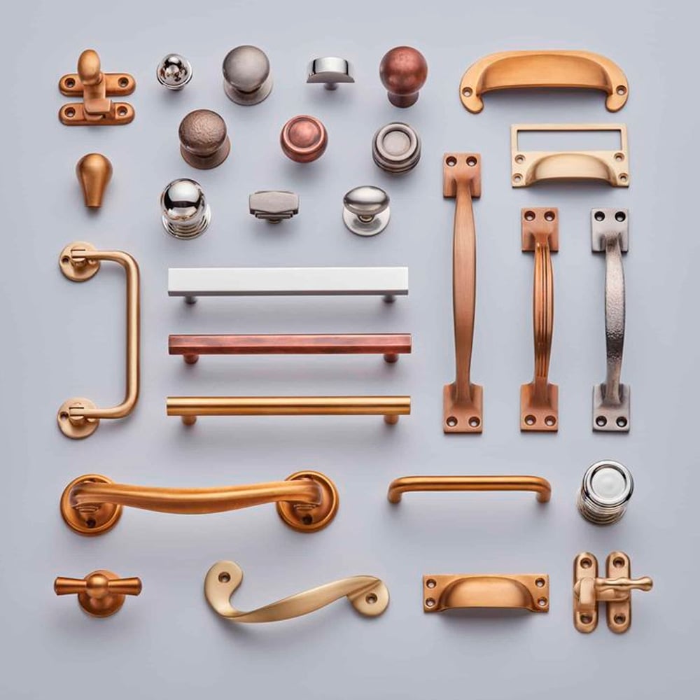 A variety of metallic cabinet handles, knobs, and pulls in different shapes, styles, and finishes, including gold, copper, silver, and bronze, are arranged neatly on a light gray background.
