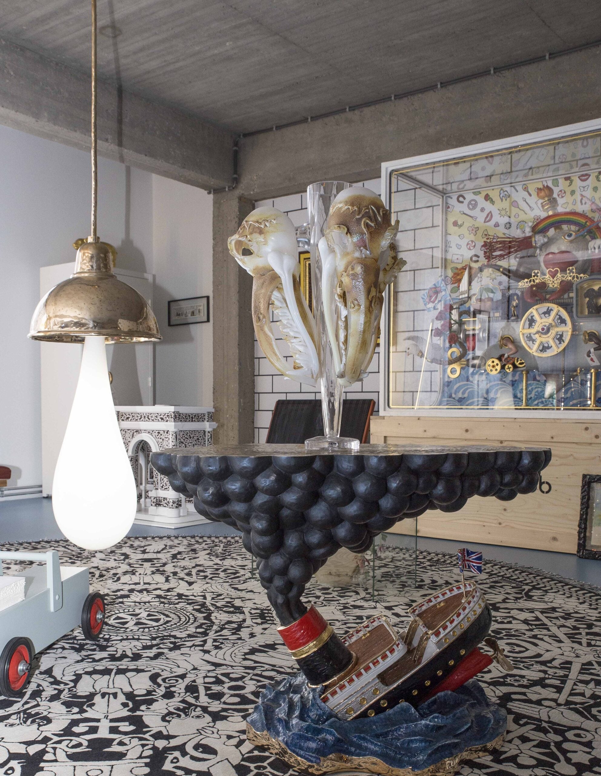 An eclectic room with a patterned floor, a table shaped like a sinking ship, two large squid sculptures, a gold hanging lamp, and shelves with various colorful objects and artwork in the background.