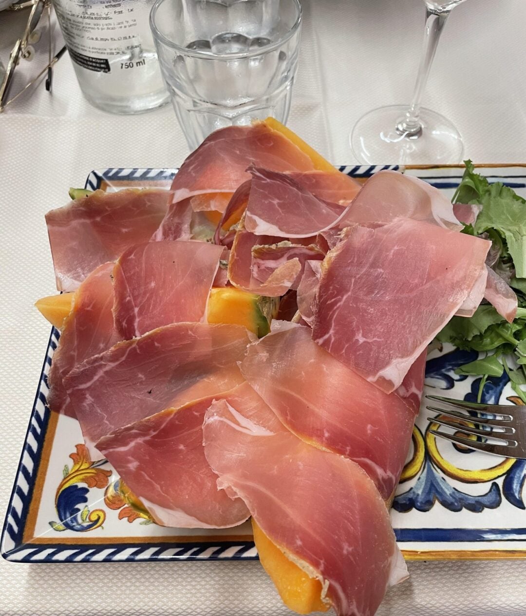 A plate with slices of prosciutto covering pieces of cantaloupe melon, garnished with a small green salad, sits on a patterned ceramic dish next to a fork, drinking glass, and a wine glass.