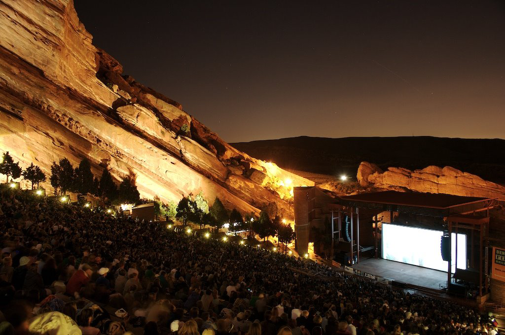 An outdoor amphitheater filled with people at night, surrounded by large illuminated red rock formations, with a bright stage at the front.