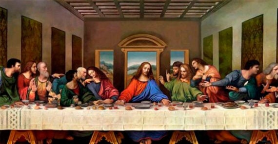 Thirteen people sit along one side of a long table covered with food, in a spacious room with three windows. The central figure is surrounded by others engaging in animated conversation.