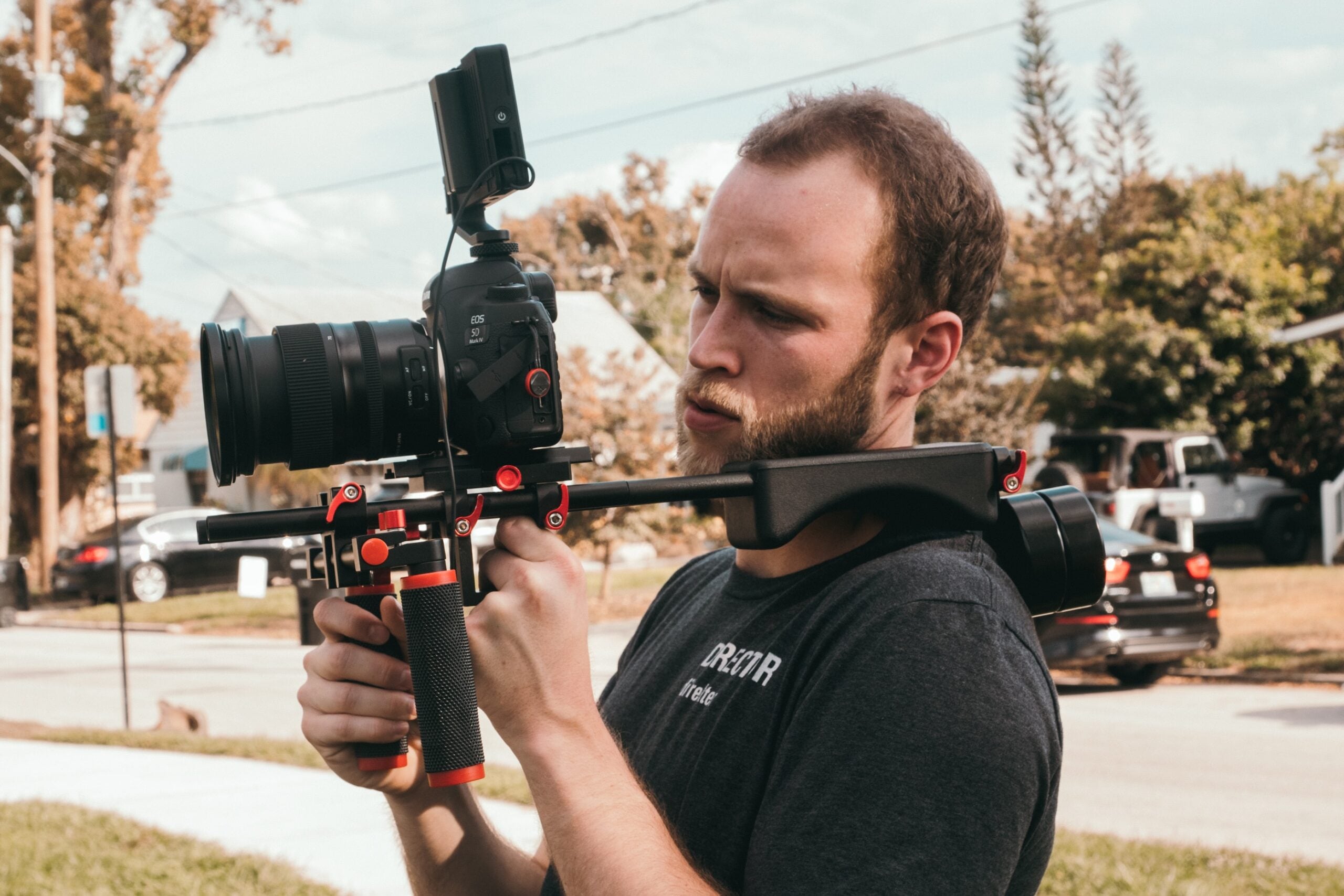 A man outdoors is filming with a professional video camera mounted on a shoulder rig, focusing intently. Trees, cars, and houses are visible in the background.
