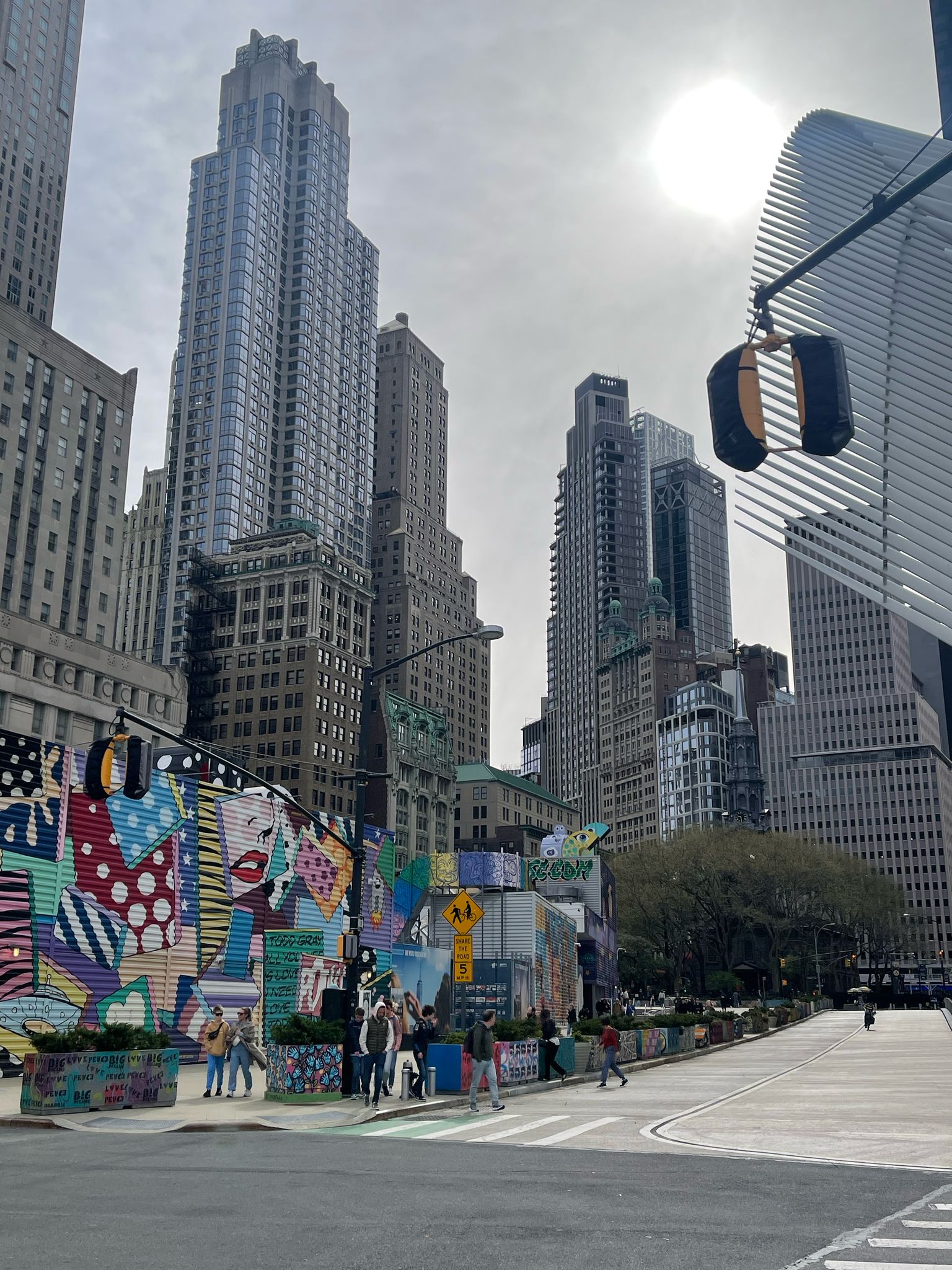 A city street scene with tall skyscrapers, colorful graffiti-covered construction barriers, pedestrians crossing, and the sun shining through a partly cloudy sky. Traffic lights and trees are also visible.