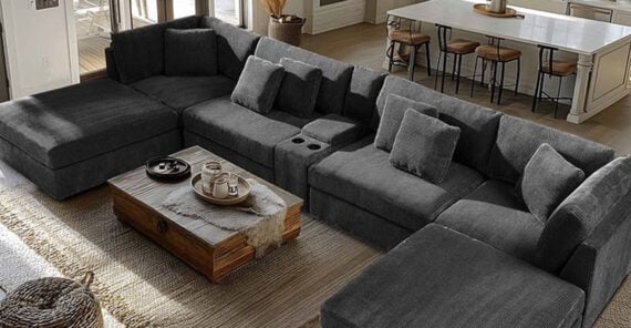 A large, dark gray sectional sofa with several cushions surrounds a wooden coffee table in a spacious living room with light wood floors and a kitchen with barstools in the background.