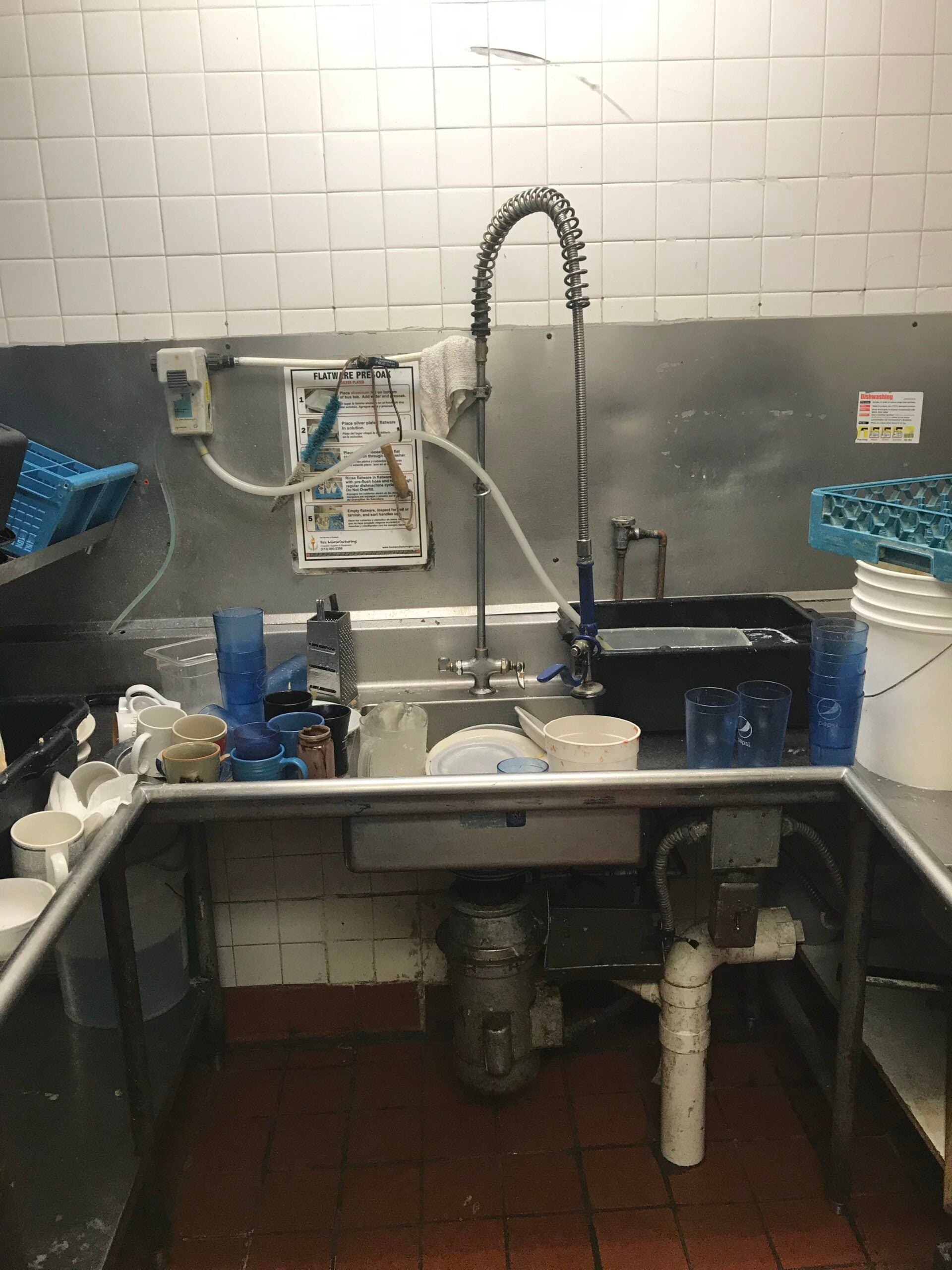 A commercial kitchen sink area with dirty dishes, cups, and utensils stacked on stainless steel countertops. A high-pressure faucet, cleaning supplies, and white tiled walls are visible.