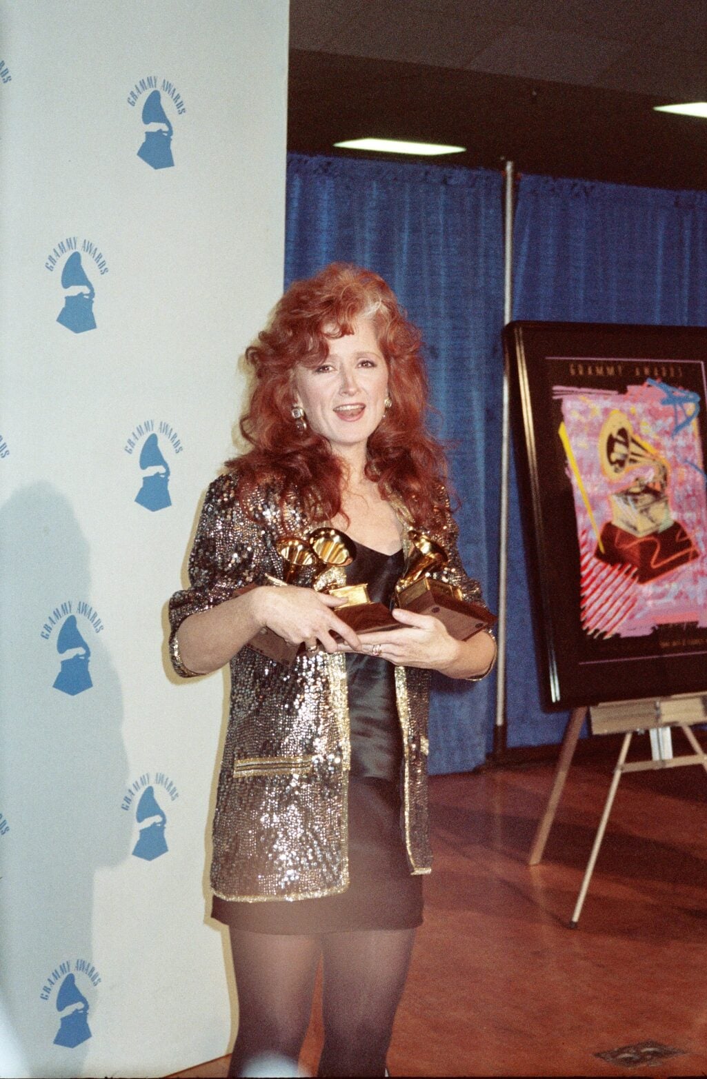 A woman with long red hair in a sparkly gold and black jacket holds an award and smiles at a Grammy Awards event, standing in front of a blue curtain and a poster.