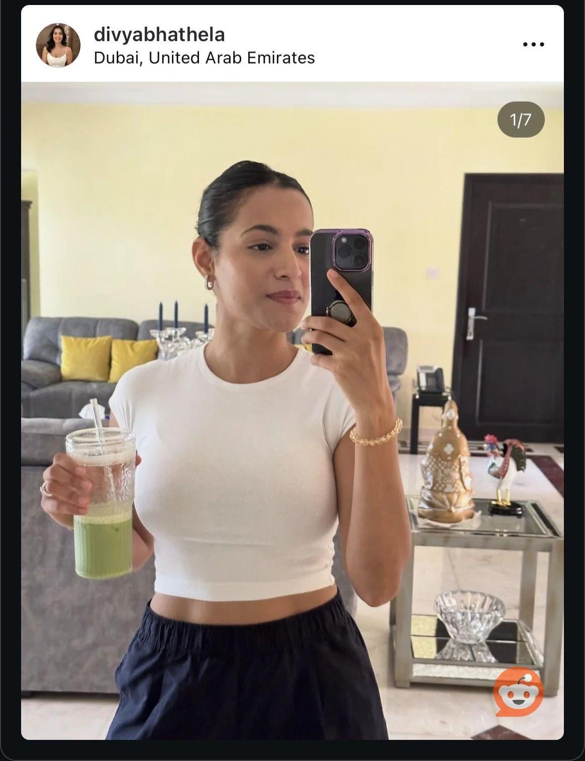 A woman in a white crop top and black pants takes a mirror selfie while holding a green drink. She stands in a modern living room with a sofa, glass table, and decorative items in the background.