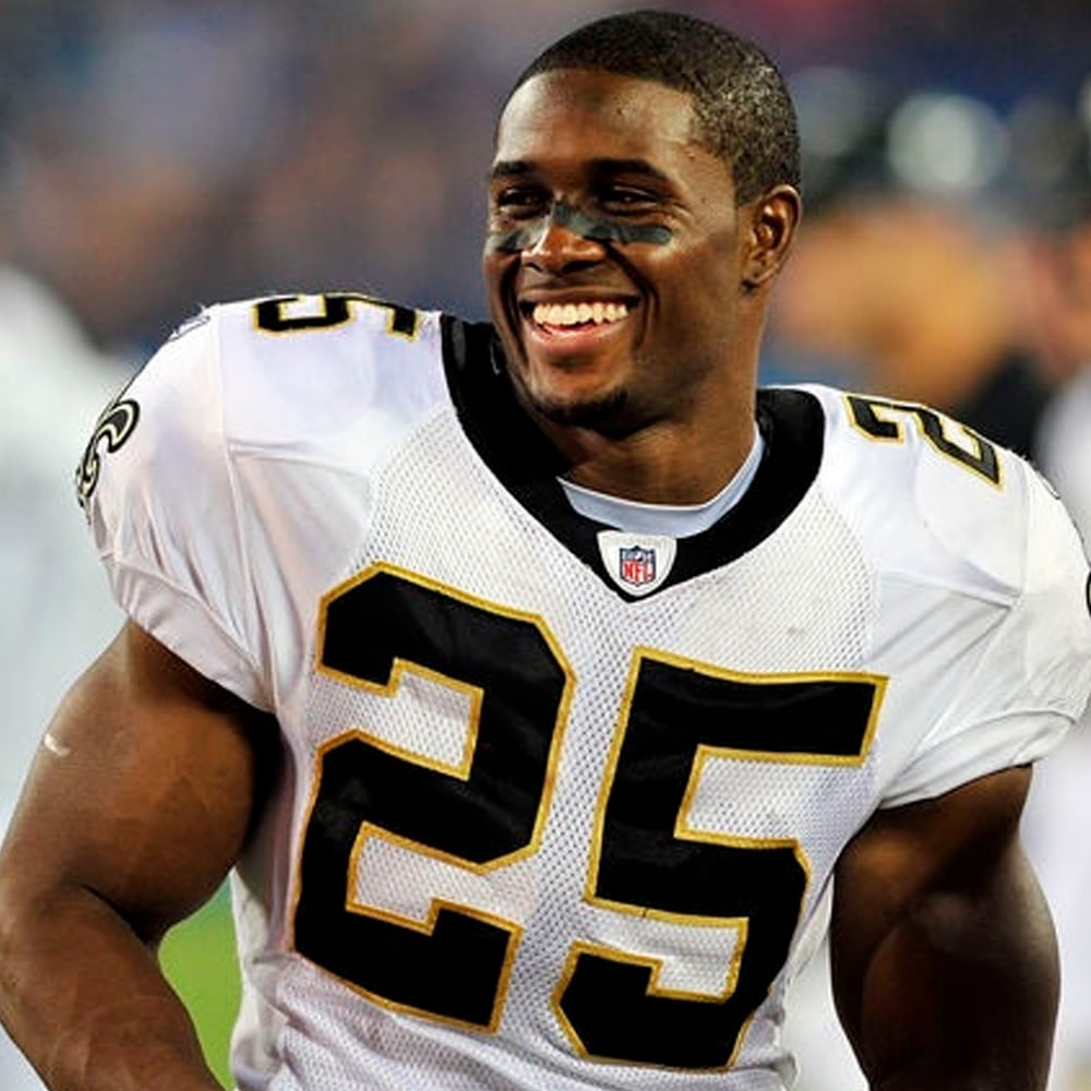 A football player wearing a white New Orleans Saints jersey with number 25 smiles on the field. He has black eye paint and is standing with his hands on his hips.