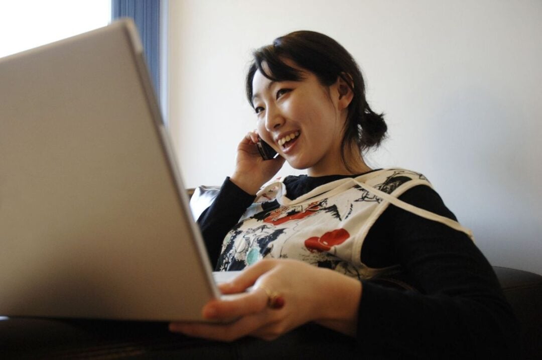 A woman sits on a couch, smiling while talking on a cellphone and using a laptop computer, wearing a patterned top with a relaxed and cheerful expression.