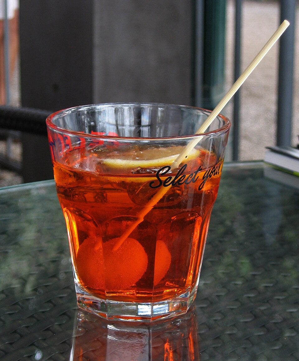 A glass of orange-colored cocktail with ice, two olives, a slice of lemon, and a straw sits on a glass table. The background is blurred.