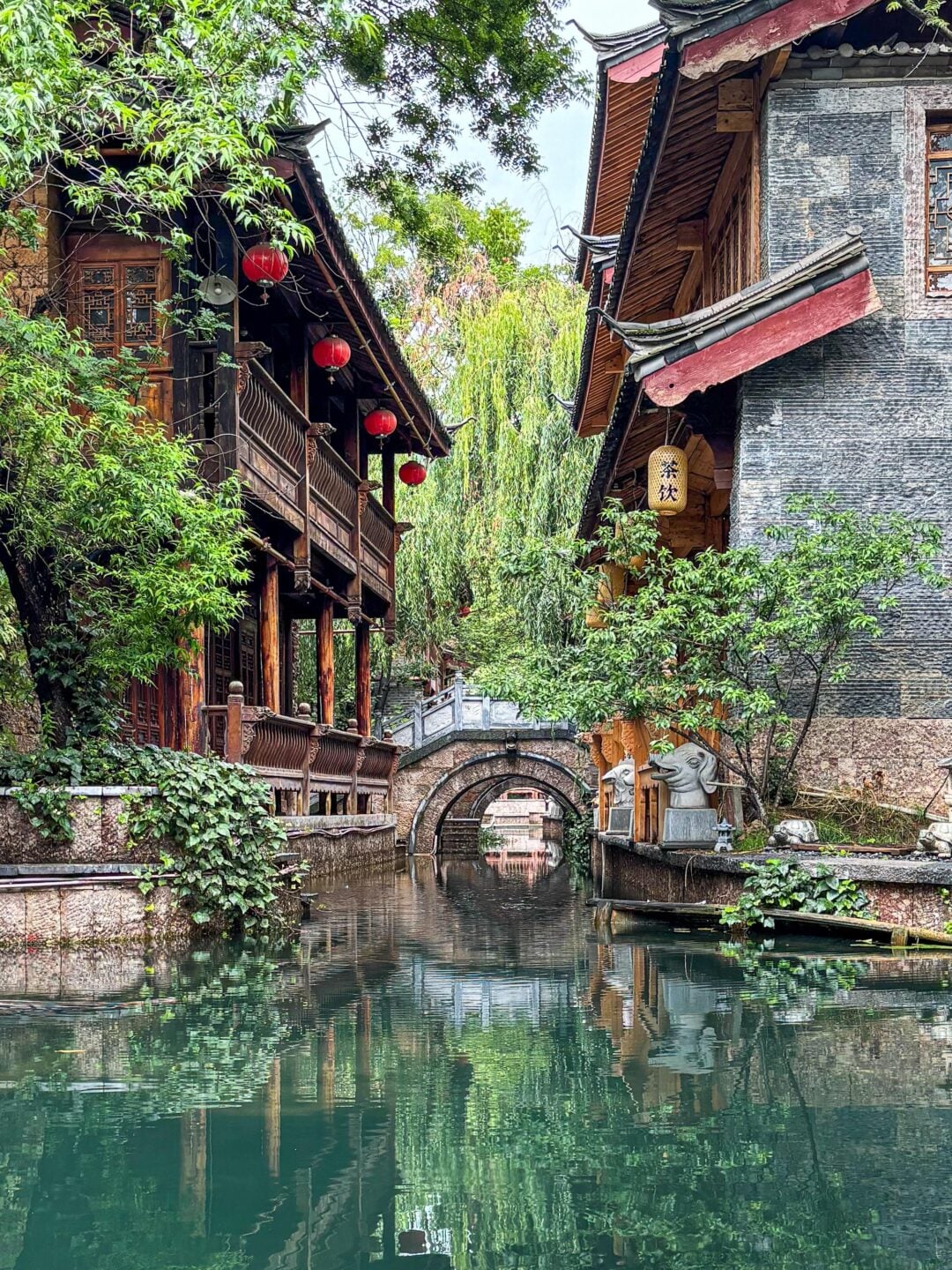 A tranquil canal lined with traditional Chinese buildings, red lanterns, lush greenery, and a stone arch bridge reflected in the calm water under a partly cloudy sky.