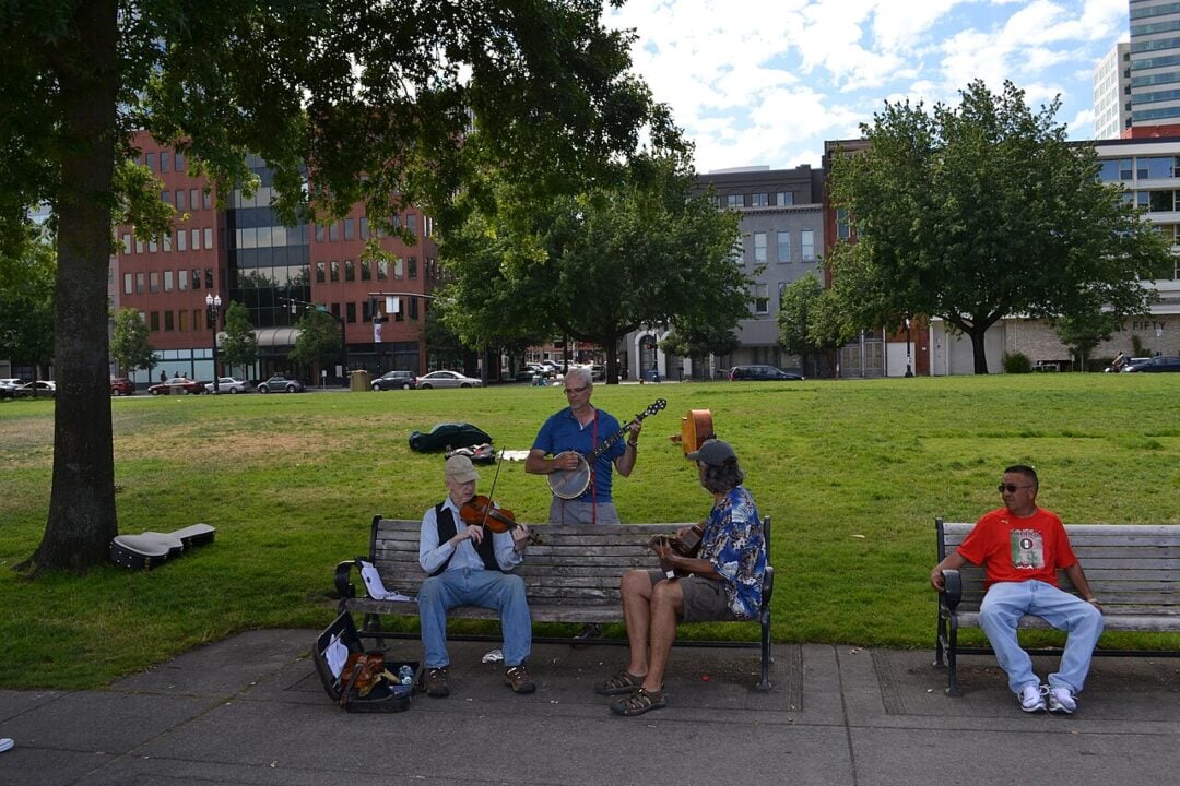Three street musicians play instruments on a sidewalk near a park, with one man sitting on a bench nearby. Trees, grass, parked cars, and buildings are visible in the background under a partly cloudy sky.