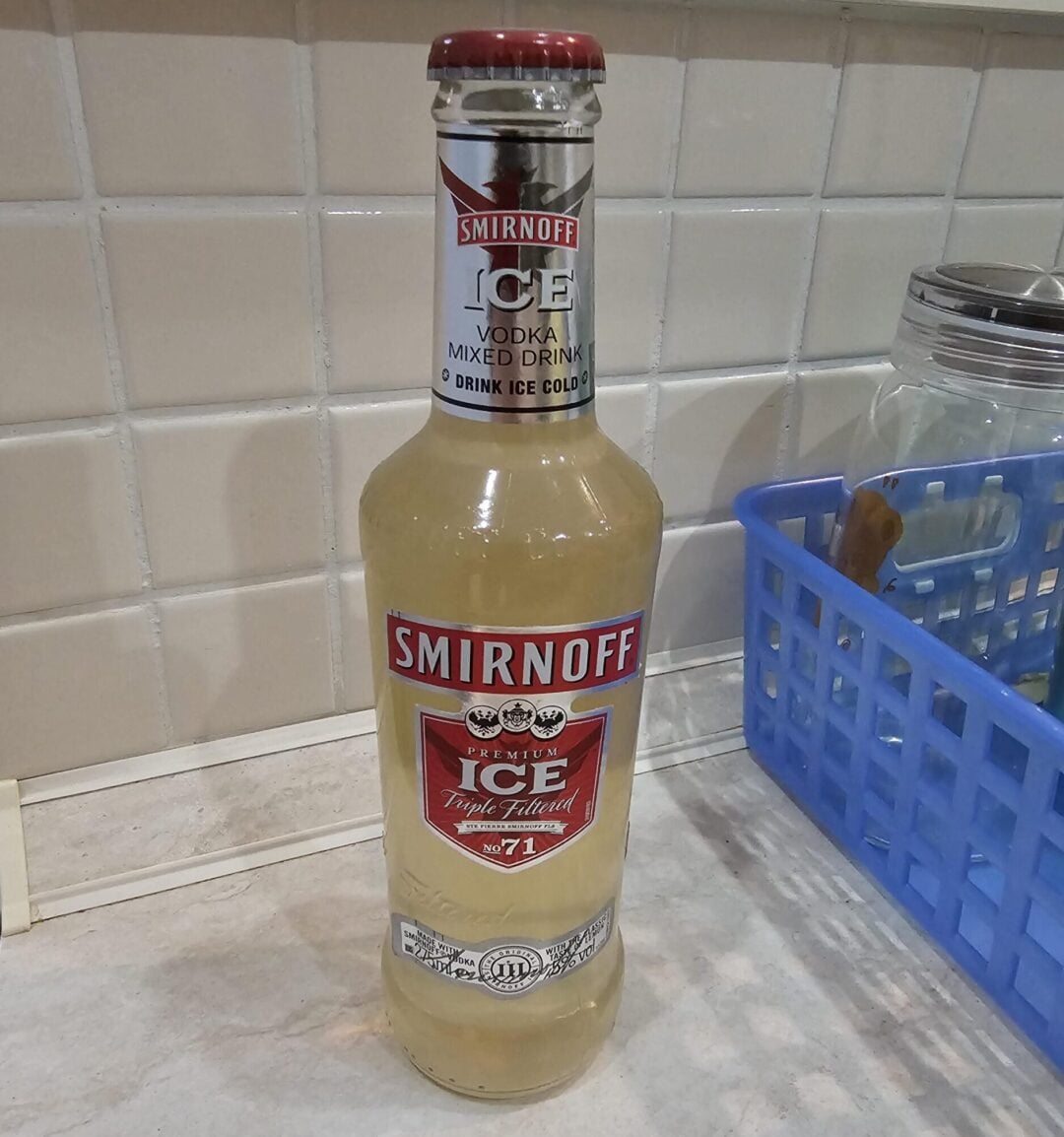 A bottle of Smirnoff Ice Triple Filtered vodka mixed drink is standing on a kitchen counter next to a blue dish rack and a glass jar, with a white tiled backsplash in the background.
