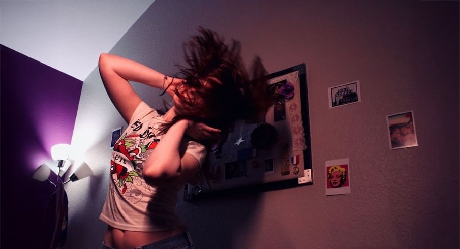 A person with long hair flips their head back, obscuring their face, in a dimly lit room with purple walls, art, and photos pinned to a board and wall behind them.