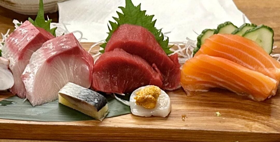 A wooden platter with assorted sashimi, including salmon, tuna, yellowtail, mackerel, and scallop, garnished with shiso leaves, sliced cucumber, and shredded daikon radish.