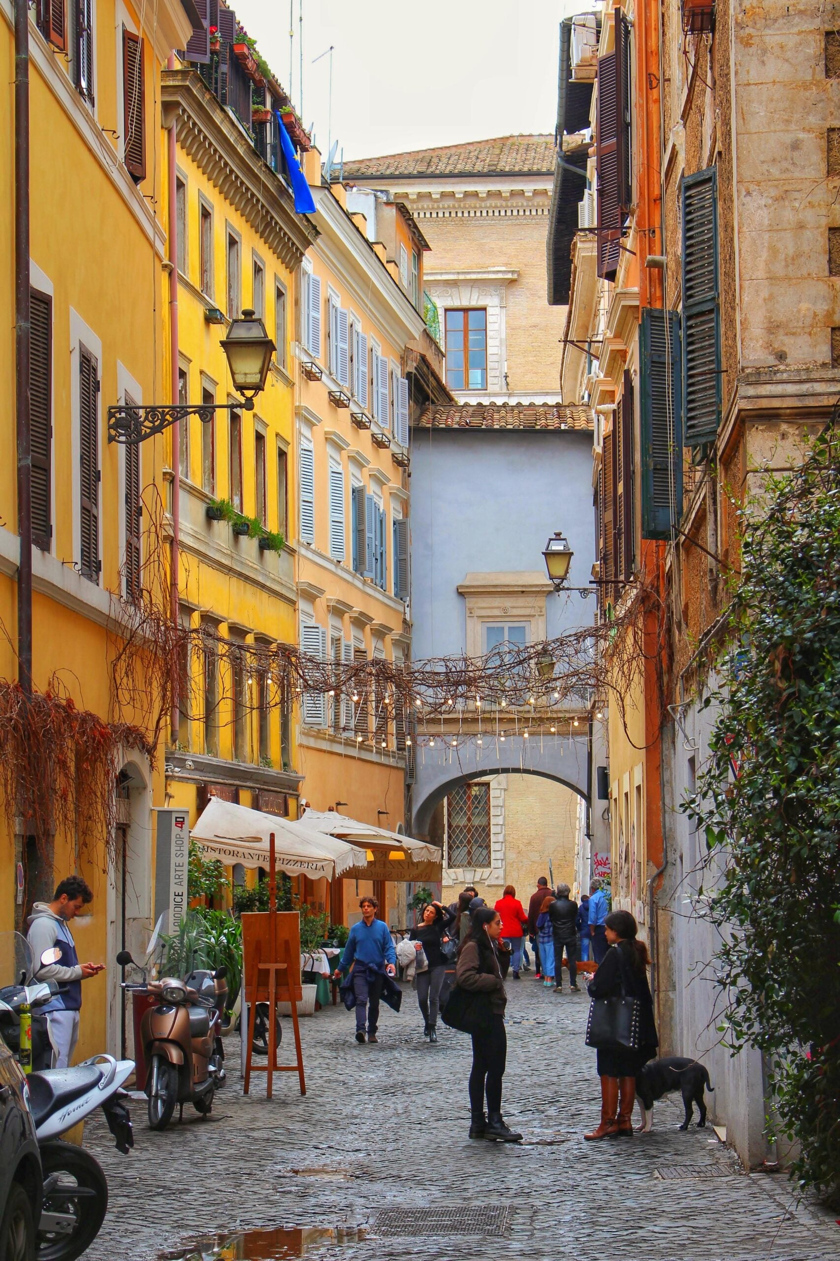 A narrow cobblestone street in a European city, lined with colorful buildings, outdoor cafes, hanging string lights, pedestrians walking, and a person with a dog in the foreground.