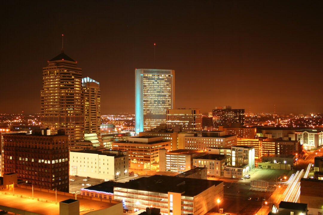 A city skyline at night with tall buildings illuminated by lights, glowing windows, and streets lit by streetlights under a dark, clear sky.
