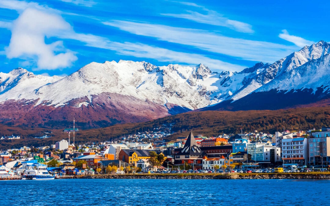 Colorful town with modern and traditional buildings sits by the water, backed by forested hills and snow-capped mountains under a bright blue sky with clouds.