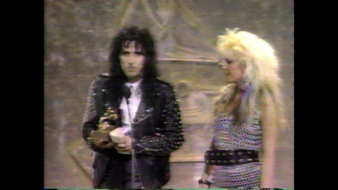 A man in a black studded leather jacket holds a trophy next to a woman with voluminous blonde hair and a studded dress. Both stand on a stage with abstract art in the background.