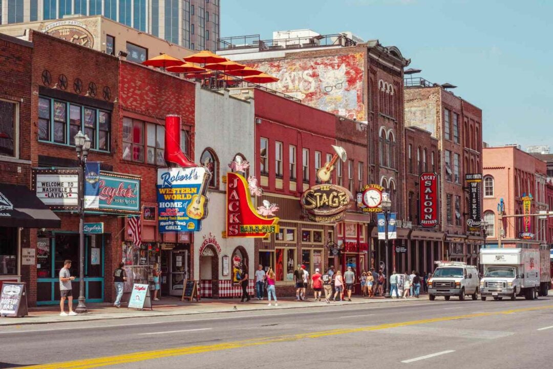 A lively street in Nashville, Tennessee, lined with colorful buildings, neon signs, bars, and restaurants. People walk on the sidewalk and two trucks are parked on the side of the road.