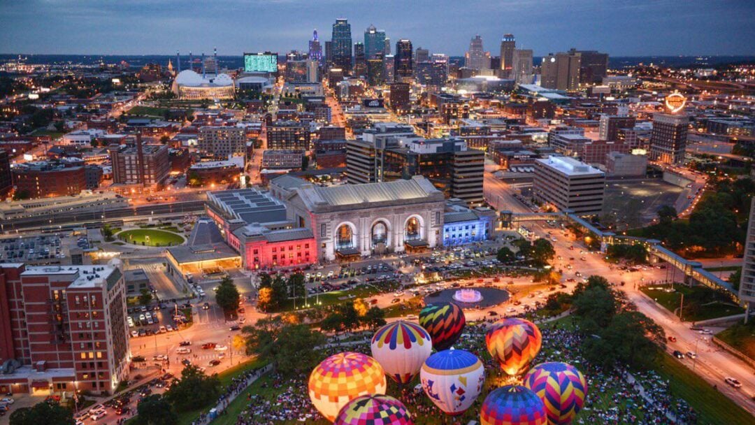 Aerial view of downtown Kansas City at dusk, with colorful hot air balloons inflated on a grassy area near Union Station, city lights illuminating the buildings and streets.