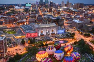 Aerial view of Kansas City at dusk, with Union Station illuminated in colorful lights and several vibrant hot air balloons glowing on the lawn in the foreground, city skyline in the background.