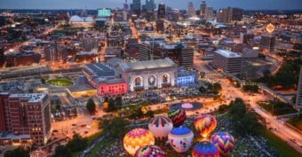 Aerial view of Kansas City at dusk, with Union Station illuminated in colorful lights and several vibrant hot air balloons glowing on the lawn in the foreground, city skyline in the background.