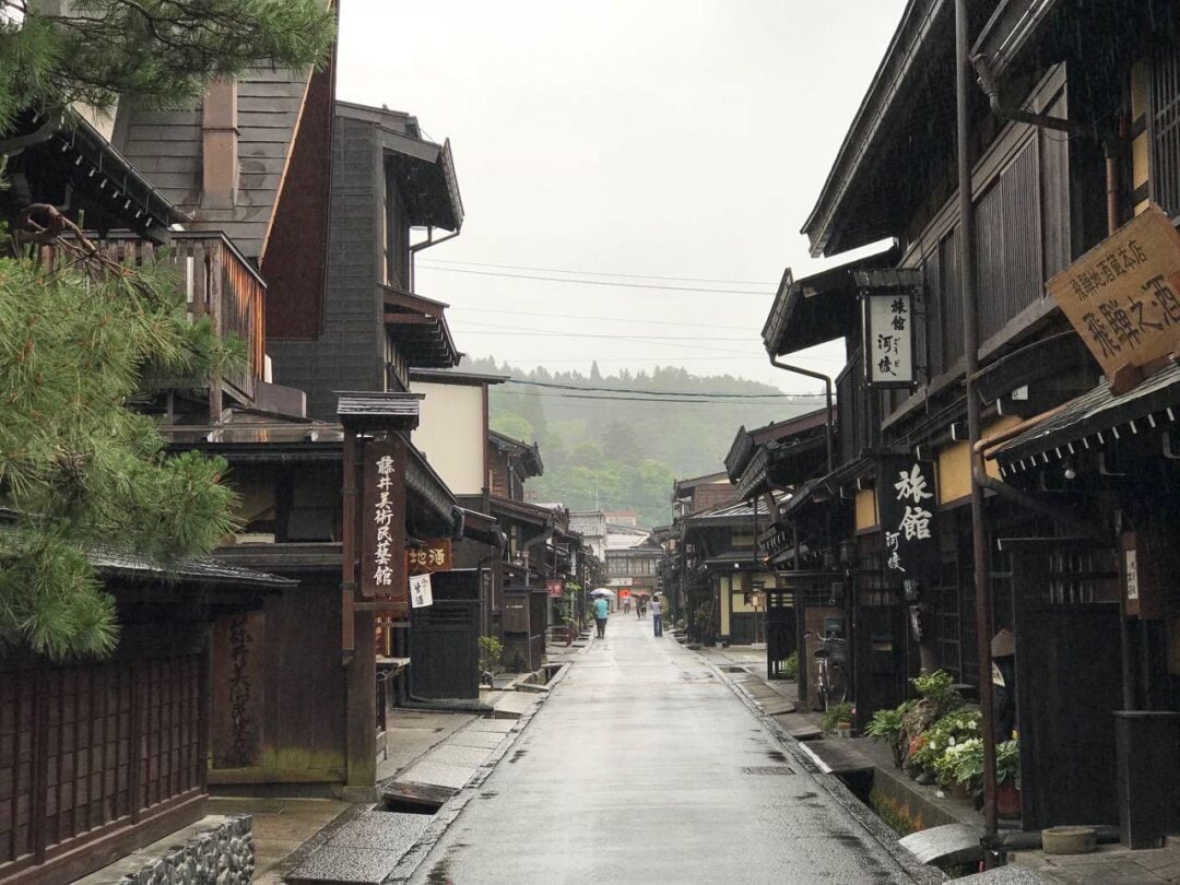 A quiet, rainy street lined with traditional wooden buildings in a historic Japanese town, with signs in Japanese and greenery in the background. The street appears empty and peaceful.