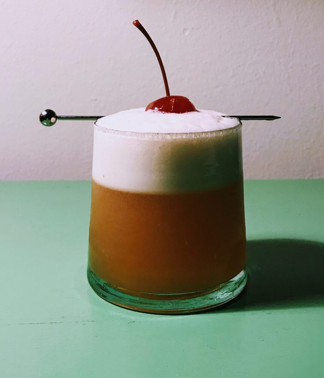 A glass of frothy whiskey sour topped with a foamy layer and garnished with a cherry on a cocktail pick, placed on a mint green surface against a plain white background.