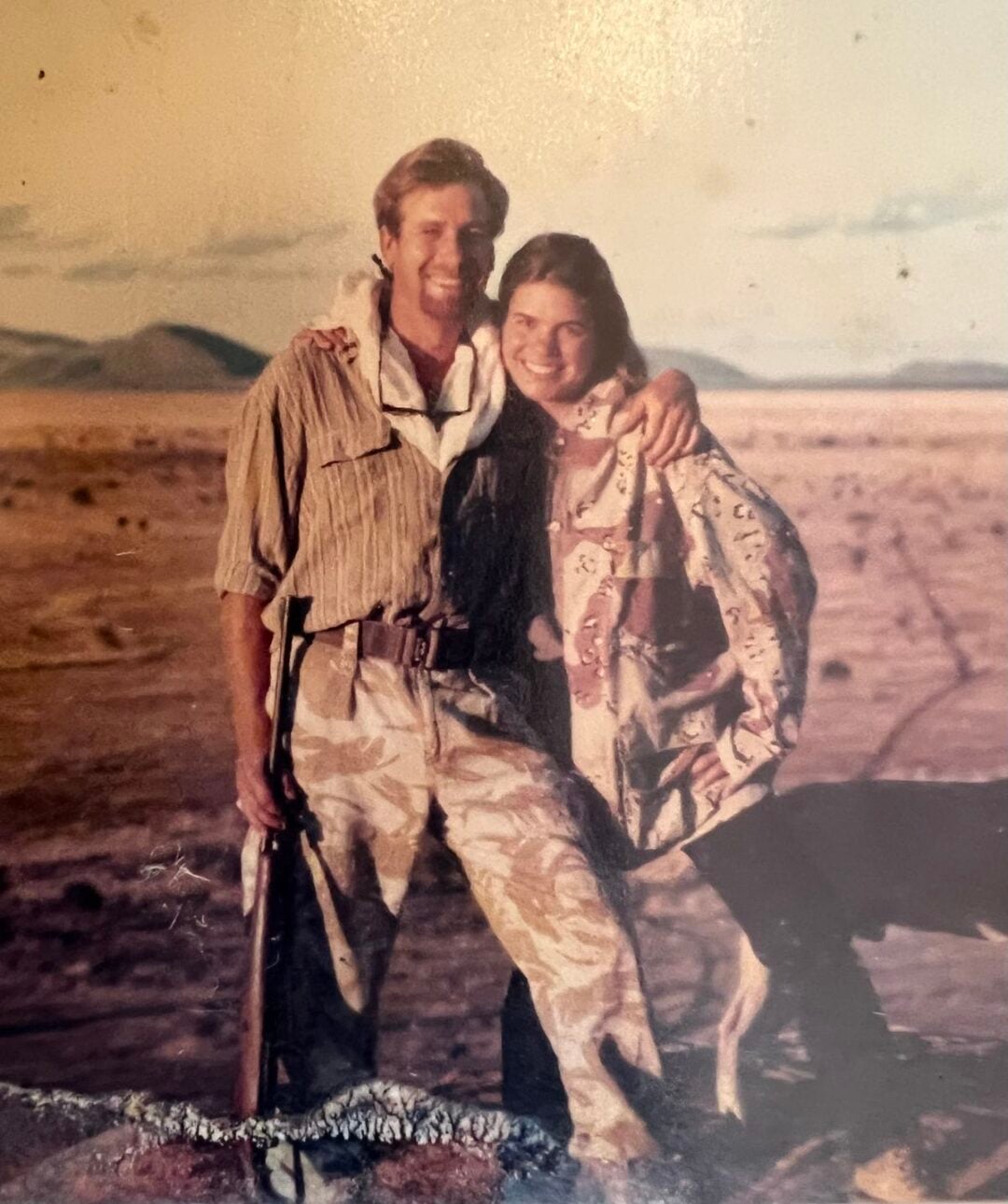 A man and woman stand close together, smiling, in a desert landscape. The man holds a rifle and wears camouflage pants. The woman, dressed in a patterned jacket, leans into him. Mountains are visible in the background, with warm lighting.