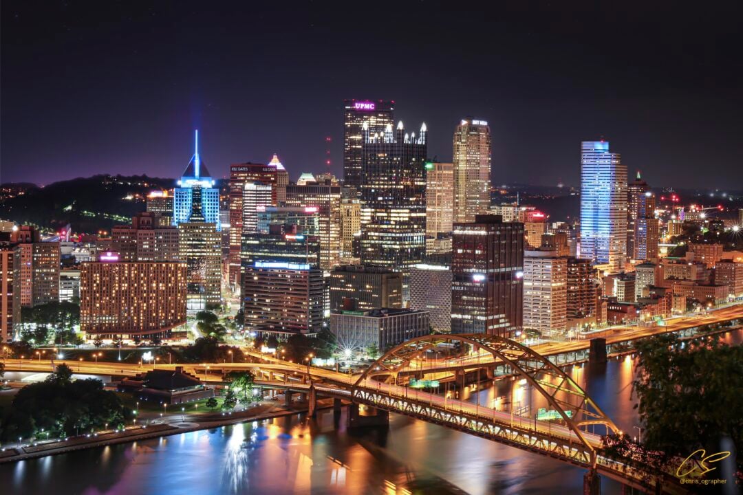 Night view of downtown Pittsburgh skyline with brightly lit skyscrapers, illuminated bridges, and the reflection of city lights on the rivers, seen from an elevated vantage point.