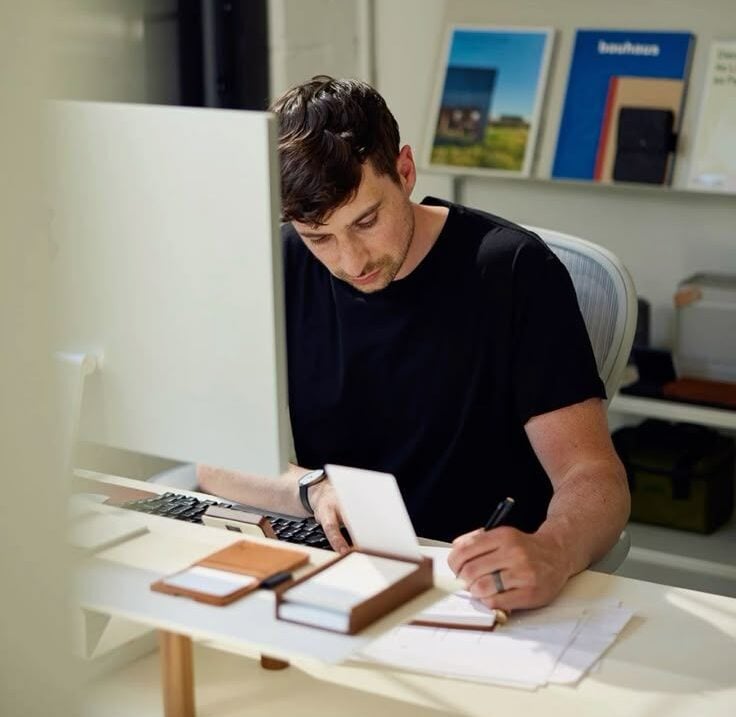 A man in a black shirt sits at a desk with a computer, writing in a notebook. The desk has note cards and papers. Books and decor items are visible on shelves in the background.