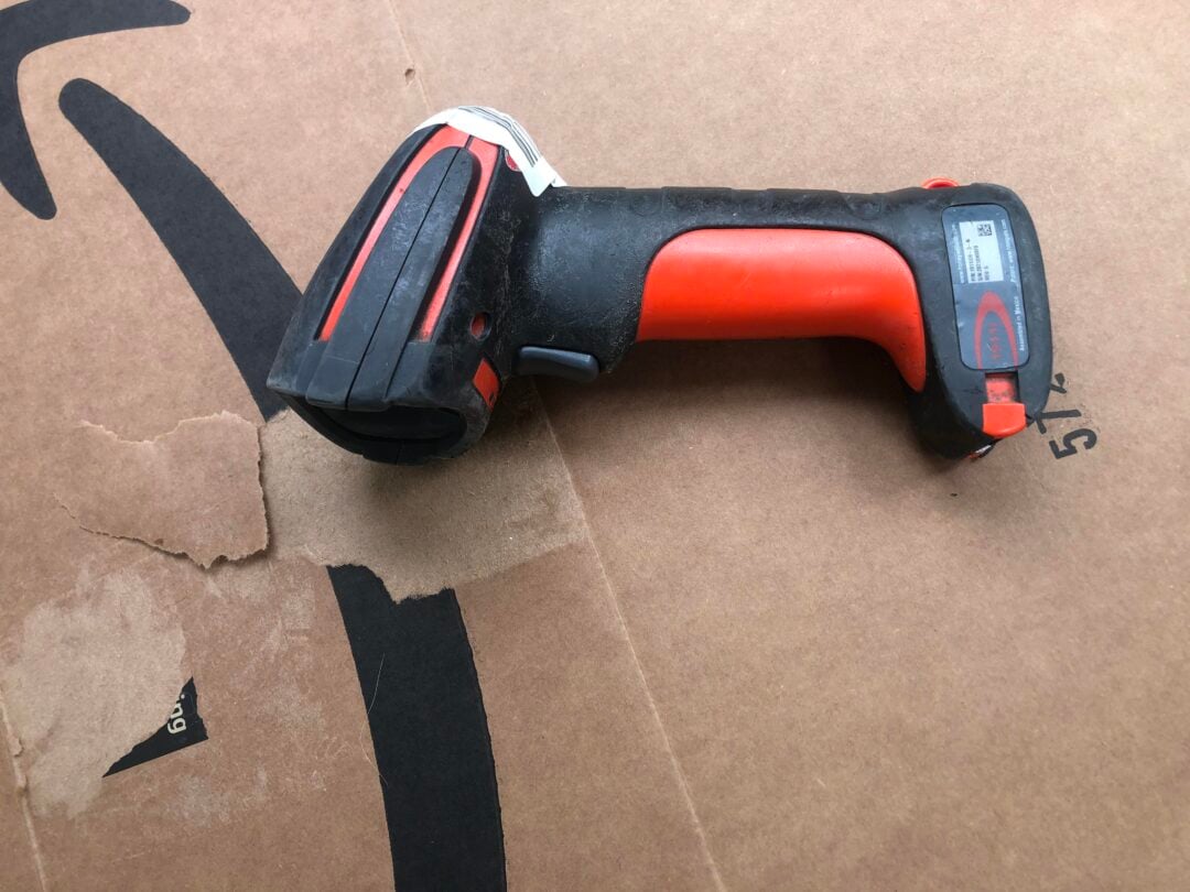 A black and red handheld barcode scanner lies on a large brown cardboard box with part of a black Amazon arrow logo visible.