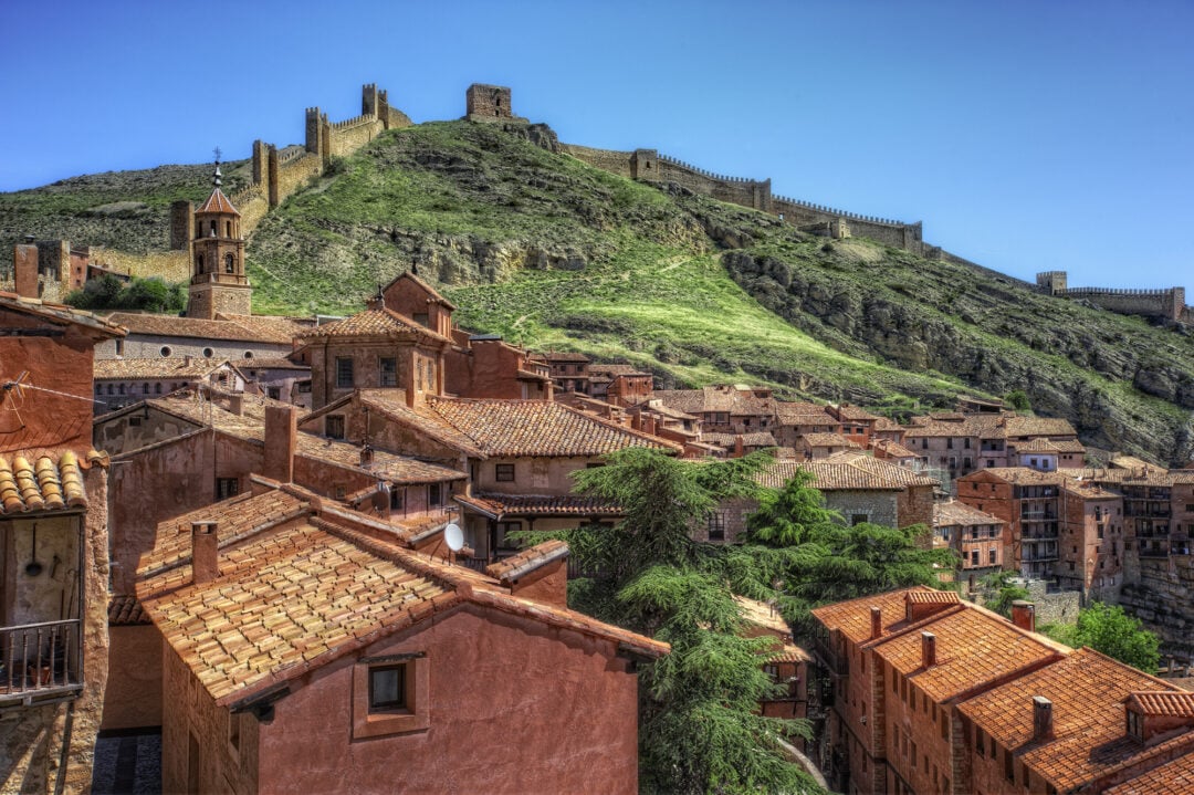 A hillside Spanish village with terracotta-roofed buildings, lush green trees, and an ancient stone fortress wall winding up a grassy hill under a clear blue sky.