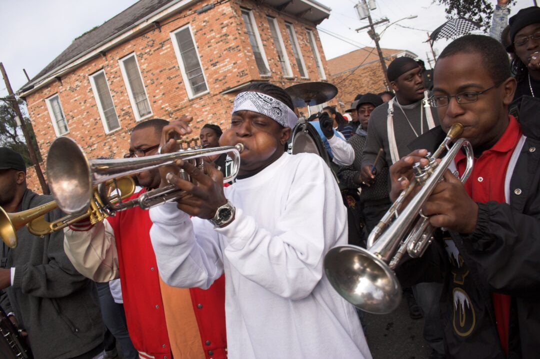 A group of musicians plays trumpets in a lively street parade. People gather around them as they perform in front of a brick building. The mood is energetic and festive.