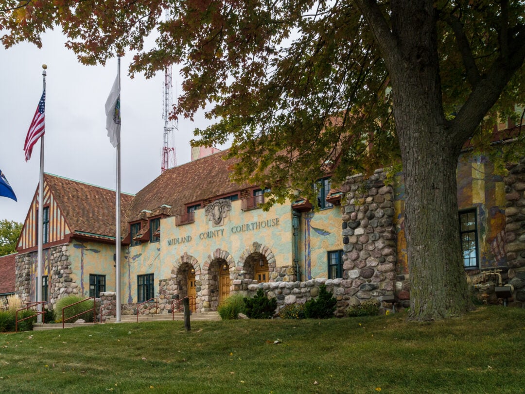 Stone and brick Midland County Courthouse with arched entrances, flags in front, and a large leafy tree partially shading the lawn on a cloudy day.