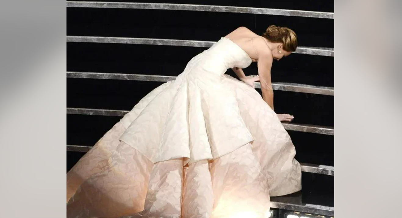 A woman in a strapless, white ball gown is kneeling on stairs, using her hands for support, as if she has just tripped or is about to get up. The background features dark steps with metallic rails.