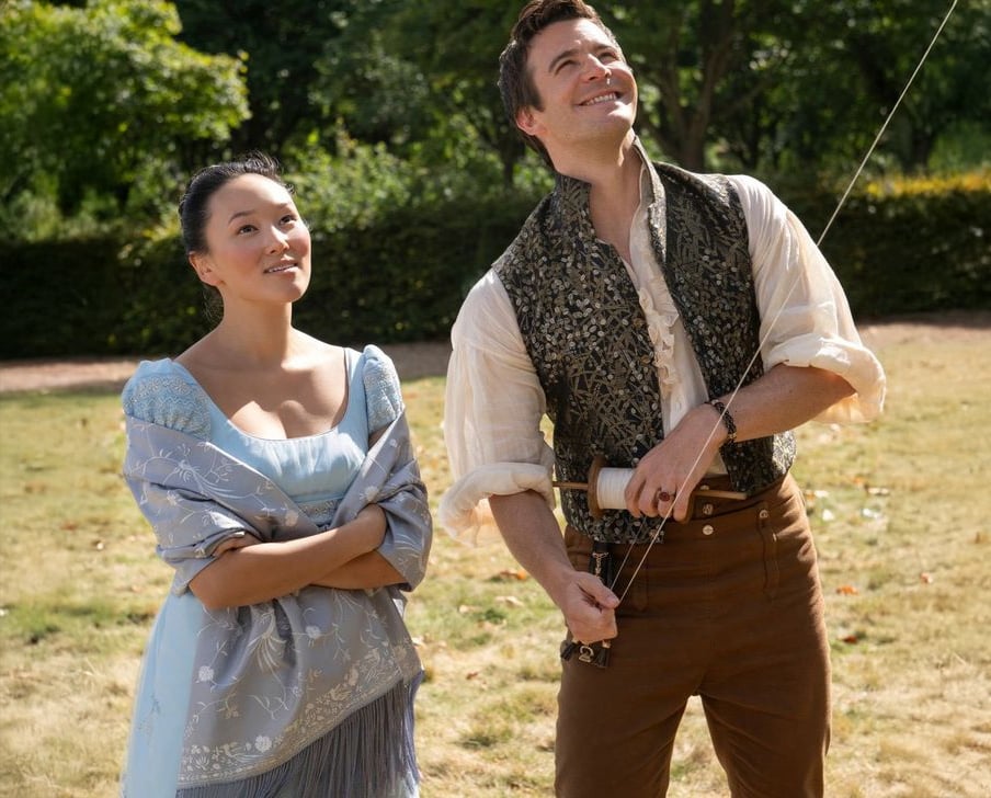 A woman in a light blue dress and a man in a vest and white shirt stand outside on grass. The man is holding a kite string and smiling upward, while the woman looks on with a soft smile. There are green trees in the background.