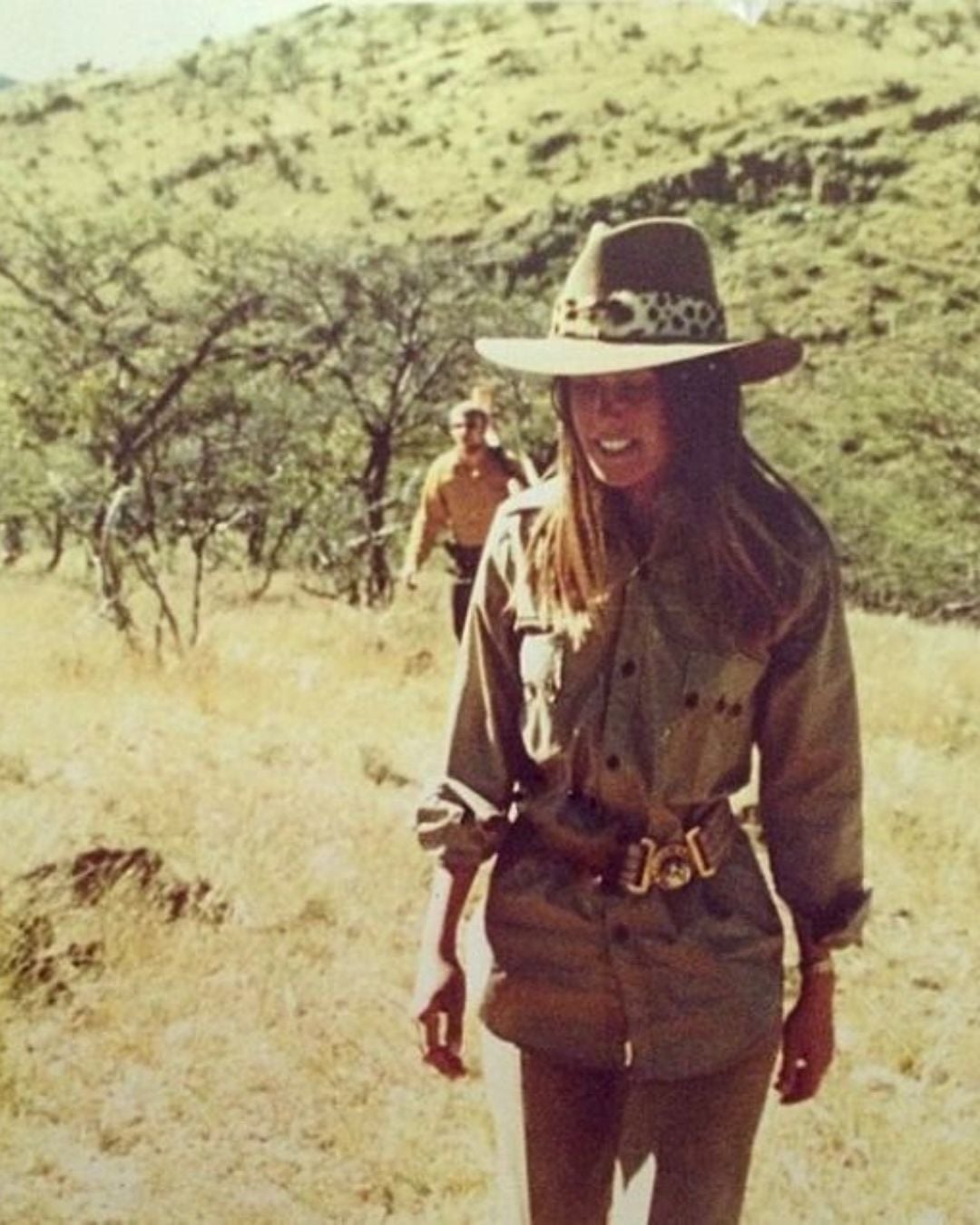 A woman wearing a wide-brimmed hat and safari-style clothing stands smiling in a grassy, hilly landscape, with a person in similar attire walking behind her among scattered trees.