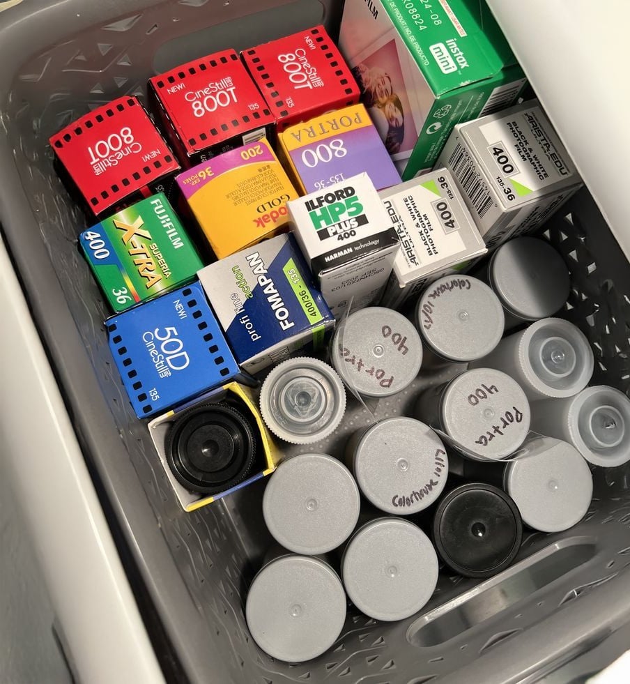 A basket filled with various film canisters and film boxes from brands like Kodak, Ilford, Fomapan, and Portra, stored neatly together in a drawer. Some canisters are labeled by hand.
