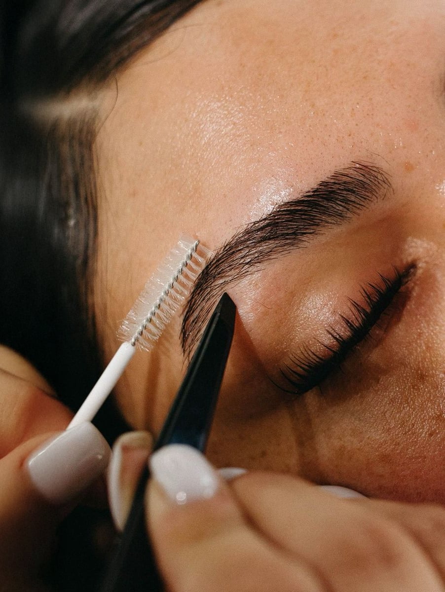 A close-up of a person's closed eye while their eyebrow is being groomed with a spoolie brush and tweezers. The skin is smooth and the eyebrow is neatly shaped.