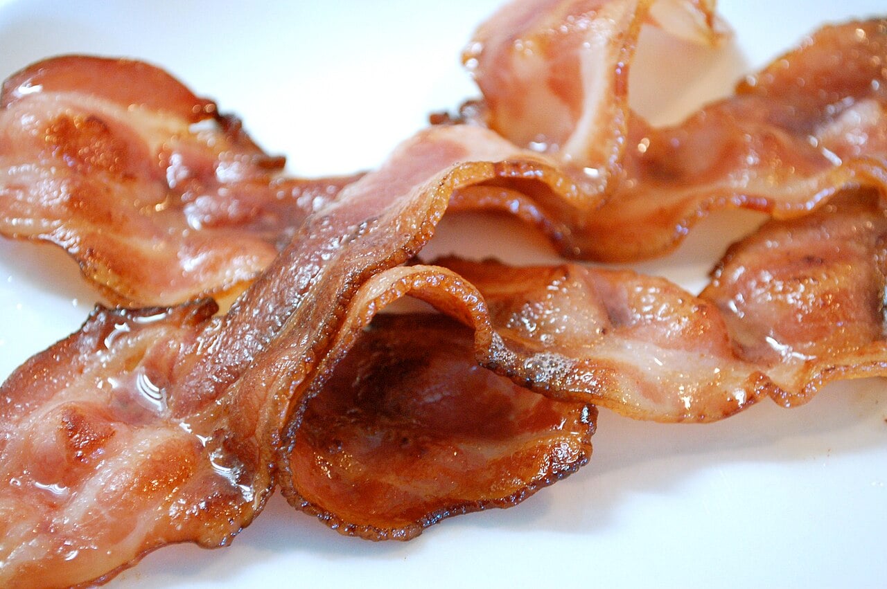 Crispy, cooked bacon strips arranged on a white plate, with a glossy, slightly oily surface and golden-brown edges.