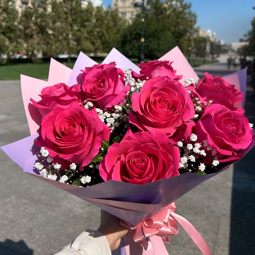 A hand holds a bouquet of vibrant pink roses with white baby's breath, wrapped in pastel pink and purple paper with a pink ribbon, outdoors on a sunny day with trees and buildings in the background.