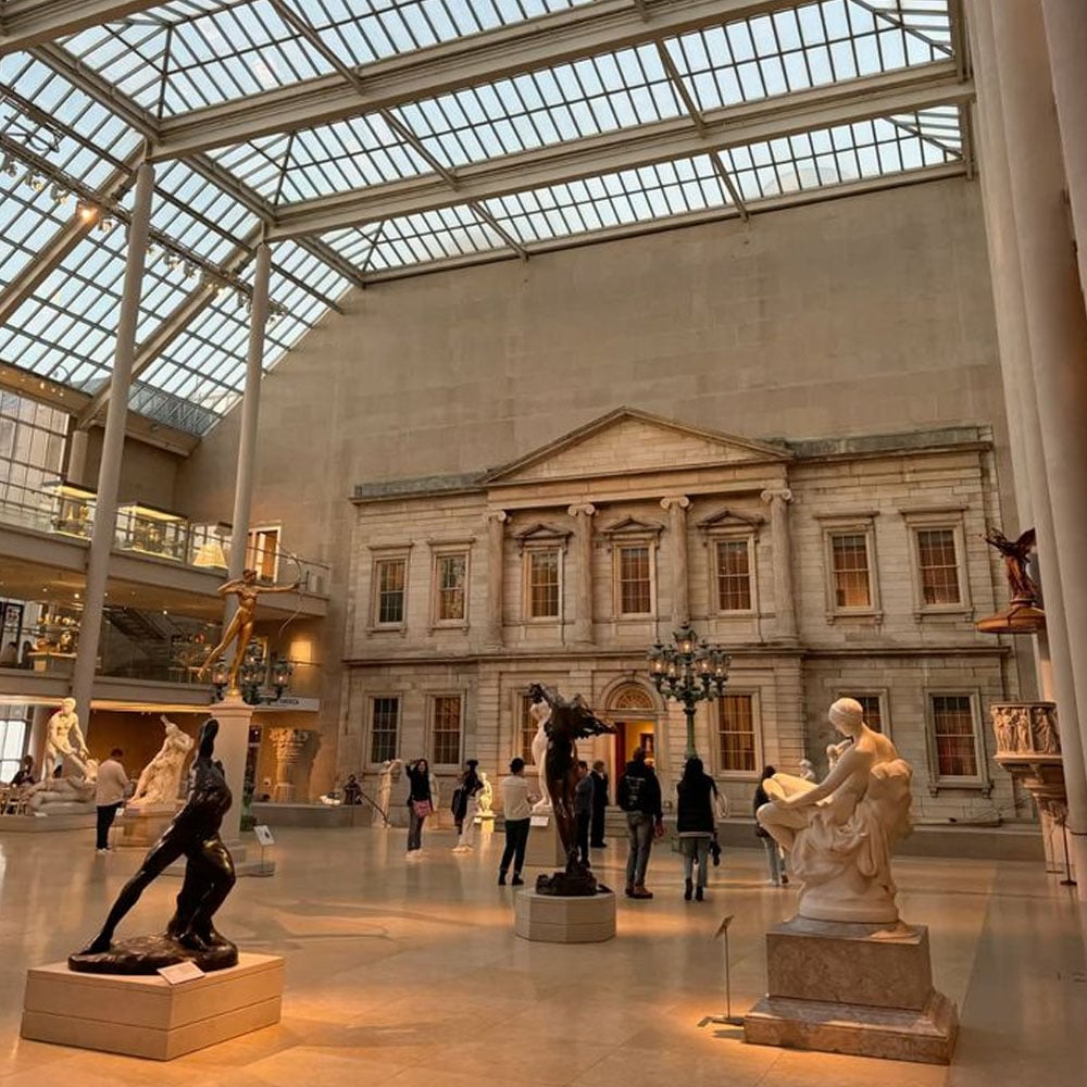 Visitors walk through a spacious museum hall with a glass ceiling, classical sculptures, and a large facade resembling a Greek or Roman building at the far end. Warm lighting highlights the artwork and architecture.
