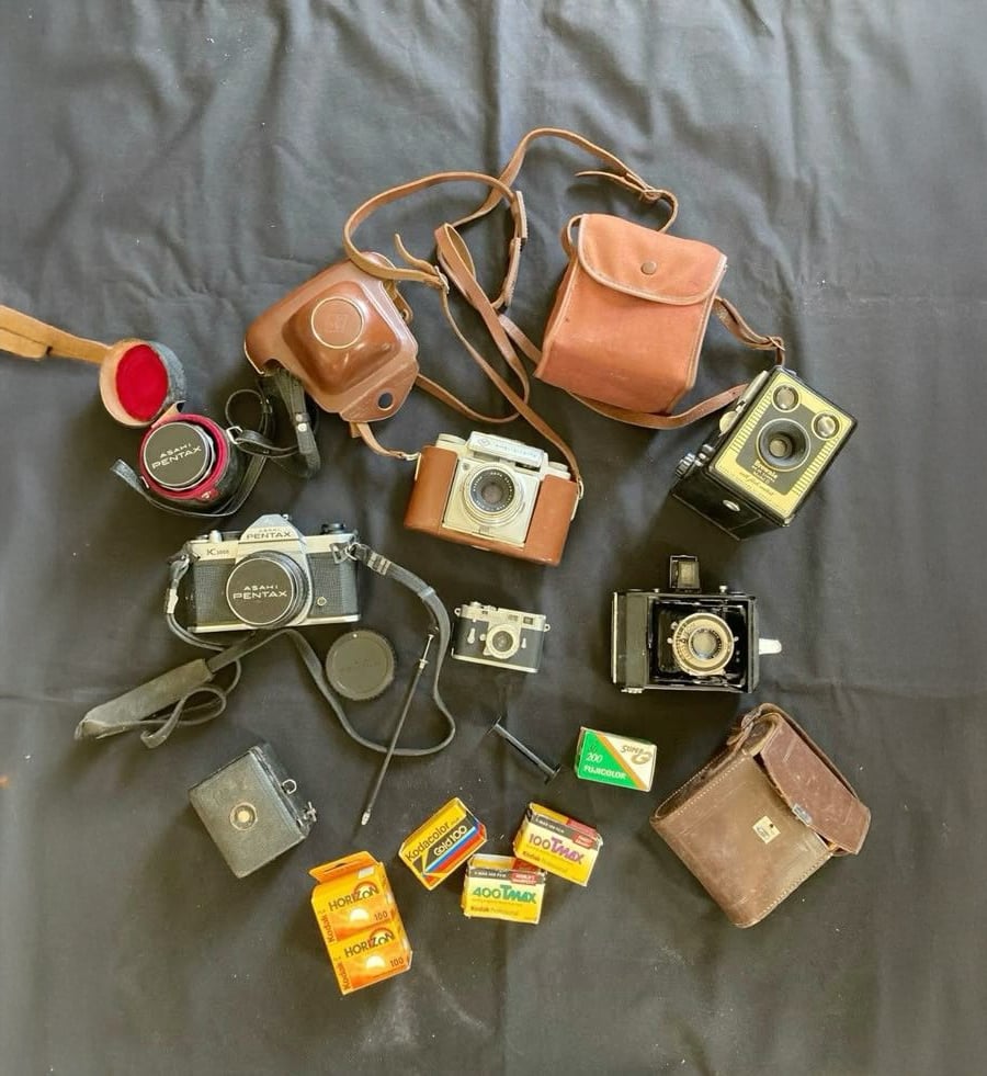 A collection of vintage cameras and film rolls arranged on a black cloth, including cameras with leather cases, several Kodak film boxes, and camera accessories.