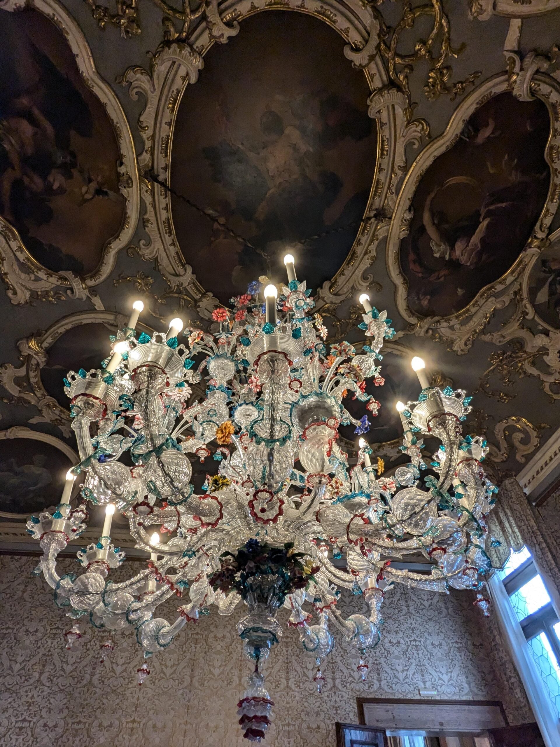 An ornate glass chandelier with colorful floral details hangs from a richly decorated ceiling featuring painted oval panels and intricate molding, in an elegant, classic room with patterned wallpaper.